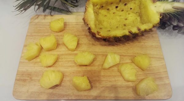 Половинка ананаса с вырезанной мякотью и кусочки фрукта на деревянной разделочной доске