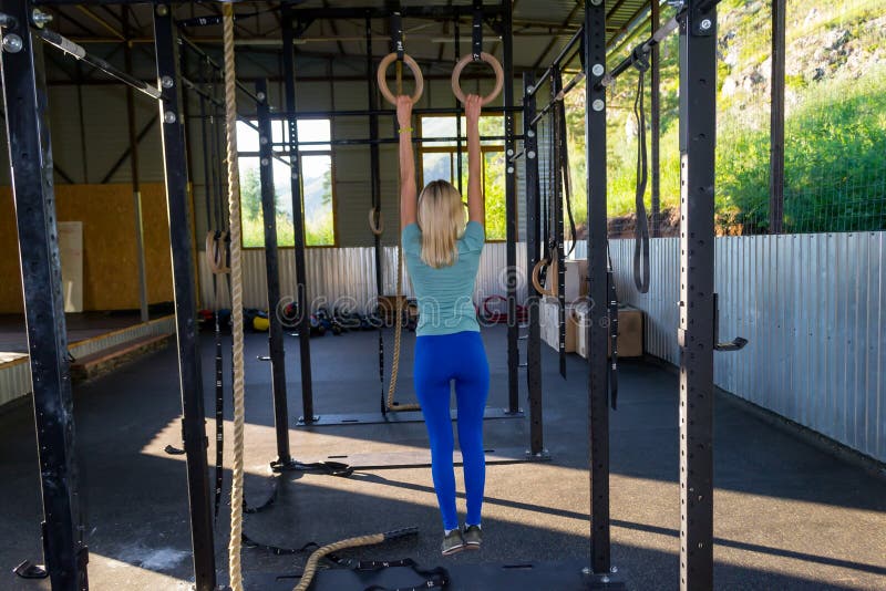 Blonde girl in blue leggings and a T-shirt from the back keeps h. Is arms outstretched for the rings aloft in an outdoor gym amidst sports equipment on the stock photo
