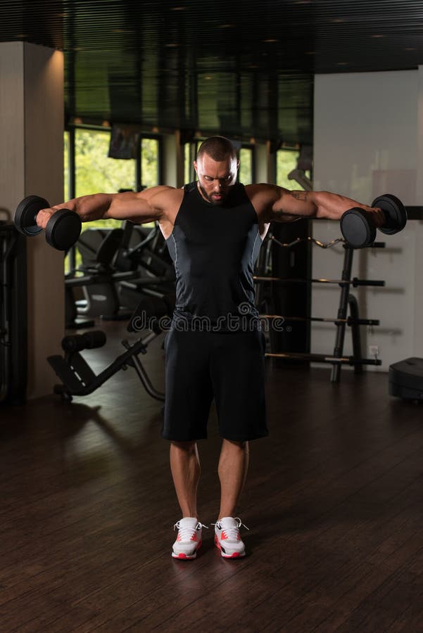 Bodybuilder Exercise Shoulders With Dumbbells. Big Man Standing Strong In The Gym And Exercising Shoulders With Dumbbells - Muscular Athletic Bodybuilder Model stock photo