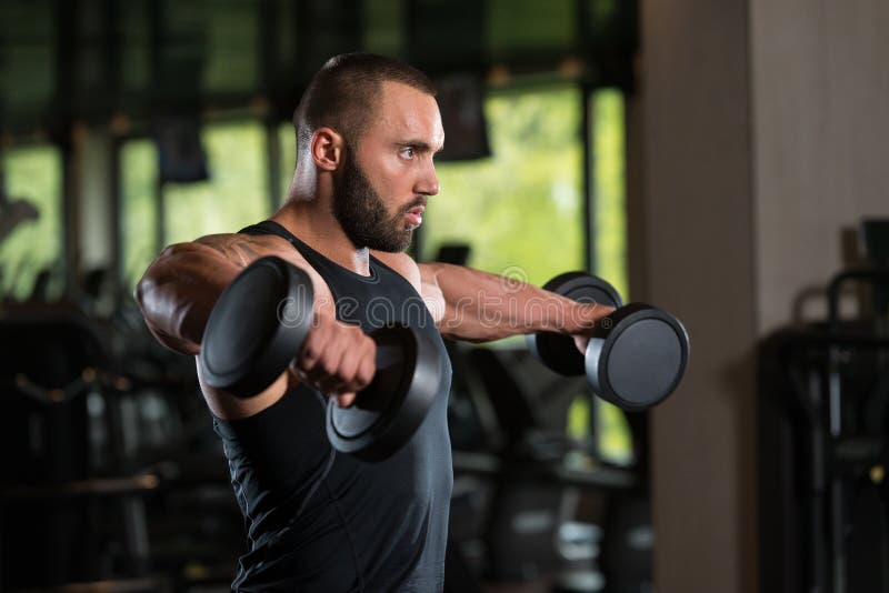 Bodybuilder Exercise Shoulders With Dumbbells. Big Man Standing Strong In The Gym And Exercising Shoulders With Dumbbells - Muscular Athletic Bodybuilder Model royalty free stock photo