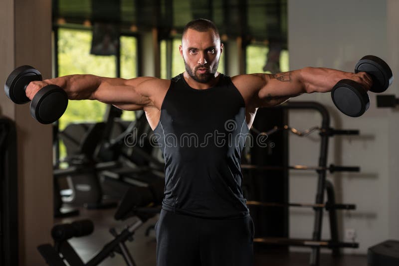 Bodybuilder Exercise Shoulders With Dumbbells. Big Man Standing Strong In The Gym And Exercising Shoulders With Dumbbells - Muscular Athletic Bodybuilder Model stock photos