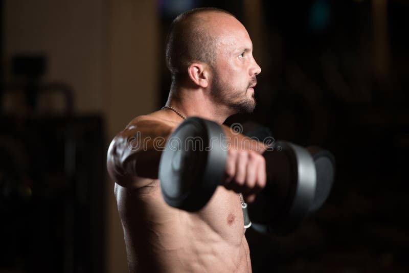 Bodybuilder Exercise Shoulders With Dumbbells. Big Man Standing Strong In The Gym And Exercising Shoulders With Dumbbells - Muscular Athletic Bodybuilder Model royalty free stock photography