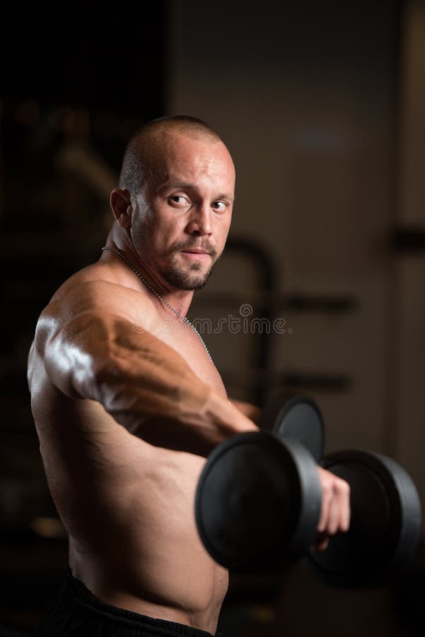 Bodybuilder Exercise Shoulders With Dumbbells. Big Man Standing Strong In The Gym And Exercising Shoulders With Dumbbells - Muscular Athletic Bodybuilder Model stock photos